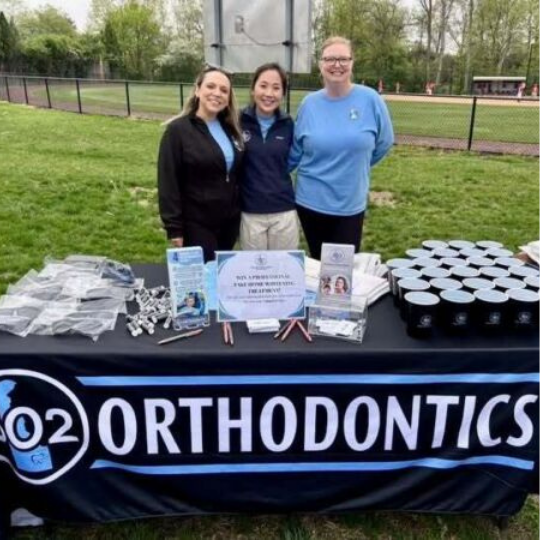Three smiling women behind a table with a 302 Orthodontics banner on a grassy sports field