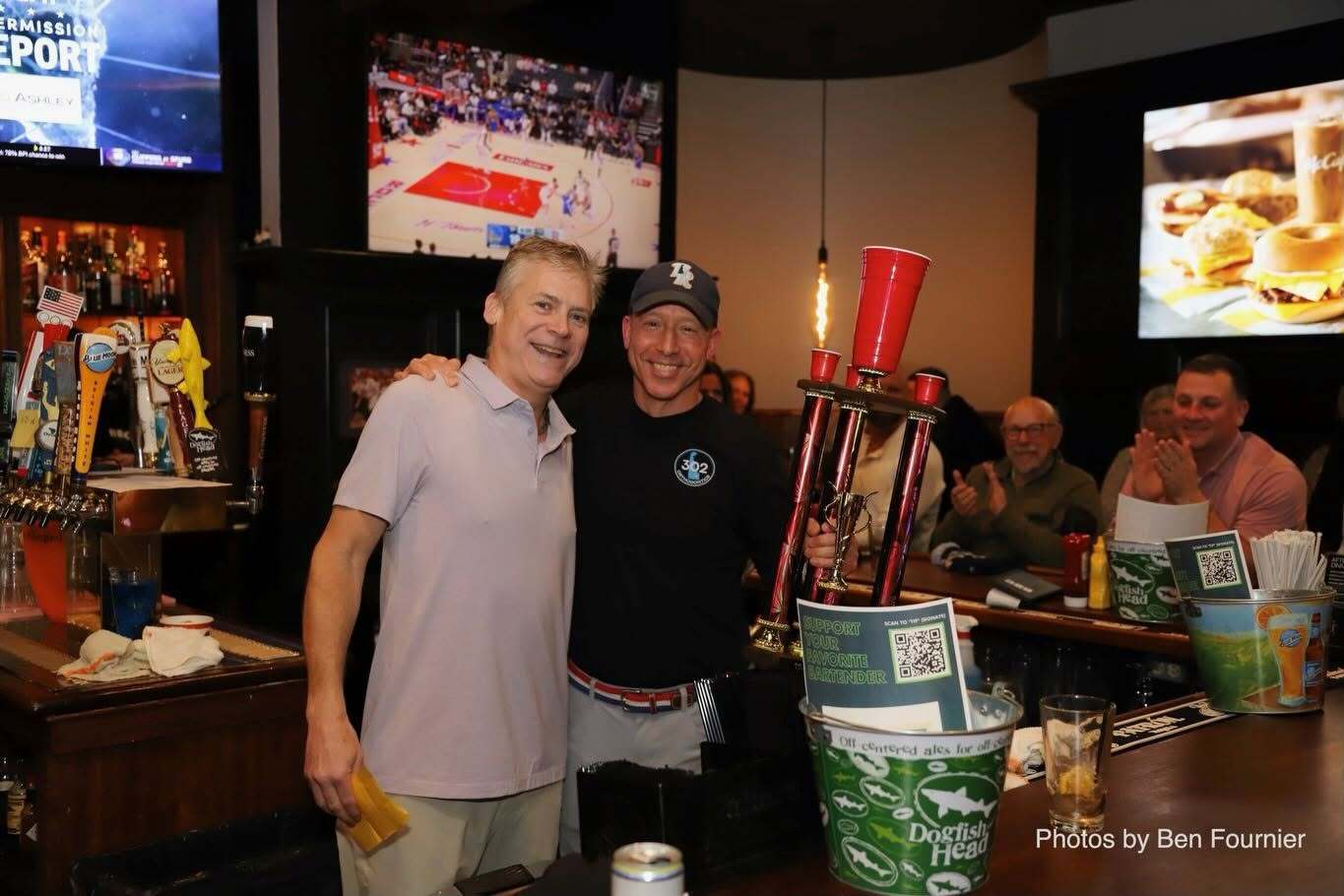 Smiling man in a cap and a black 302 logo shirt holding a tall trophy beside another smiling man at a bar