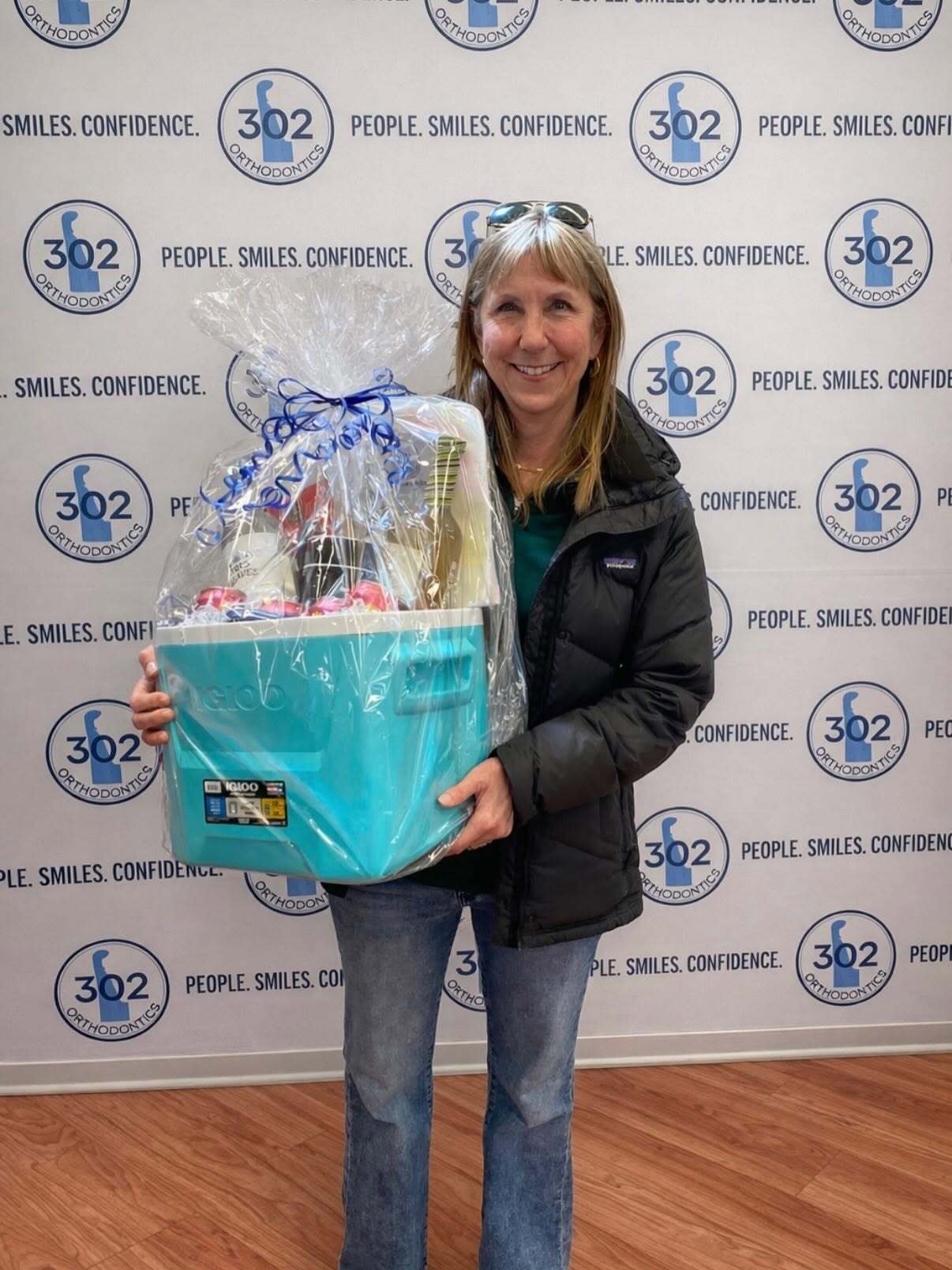 Smiling woman holding a cellophane-wrapped teal Igloo cooler with a bow, in front of a 302 Orthodontics logo backdrop