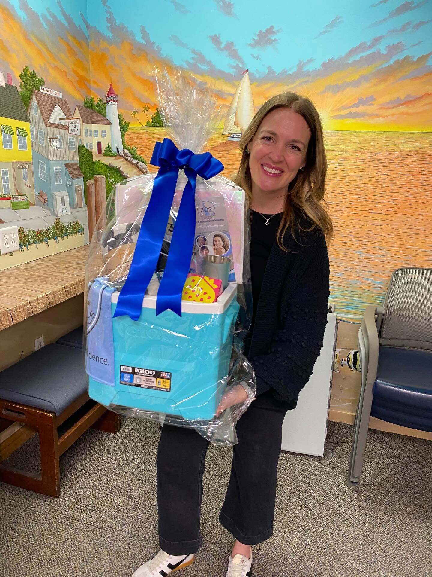 Smiling woman holding a cellophane-wrapped teal Igloo cooler with a blue bow in front of a painted harbor mural